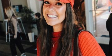 A smiling woman holds a coffee cup and wears a hat.