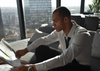 Young professional reading a newspaper in an urban office with cityscape view.