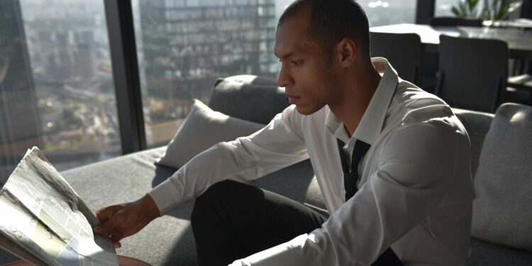 Young professional reading a newspaper in an urban office with cityscape view.